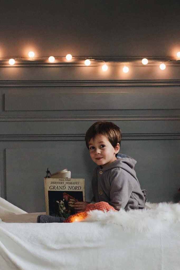 petit garçon futur grand frère souriant lit un livre sur son lit bohème avec guirlande lumineuse
