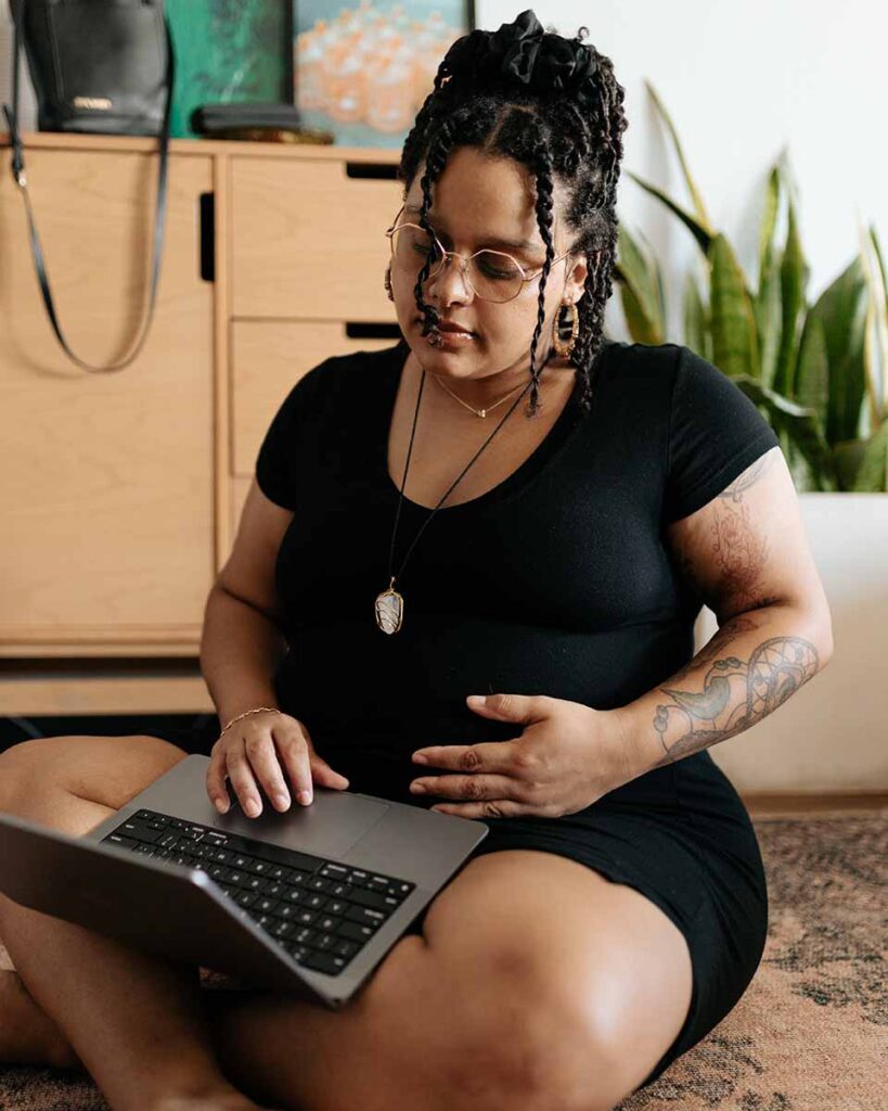 femme métis enceinte assise en tailleur par terre avec ordinateur sur les genoux robe noire et pendentif 