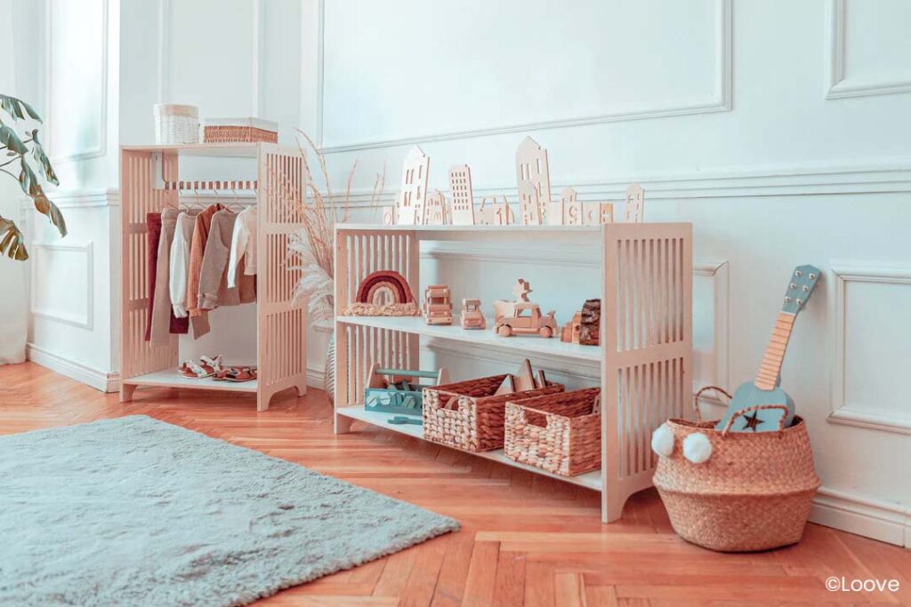 Photographie d'une chambre d'enfant aménagée avec soin selon la pédagogie Montessori. Au centre, une étagère en bois clair avec des compartiments présente des jouets en bois tels qu'une mini ville avec ses bâtiments, des véhicules et un arc-en-ciel empilable. À gauche, une armoire ouverte dévoile des vêtements de bébé suspendus à des cintres, avec un espace en dessous pour ranger des chaussures. Des plumes décoratives émergent d'une fente de l'étagère, ajoutant une touche naturelle. À droite, une petite guitare bleue est posée contre le mur à côté d'un panier contenant des jouets doux. Le sol est en parquet avec un tapis doux en premier plan. La pièce, baignée d'une lumière douce, inspire la tranquillité et l'éveil sensoriel, favorisant l'autonomie de l'enfant à la maison.