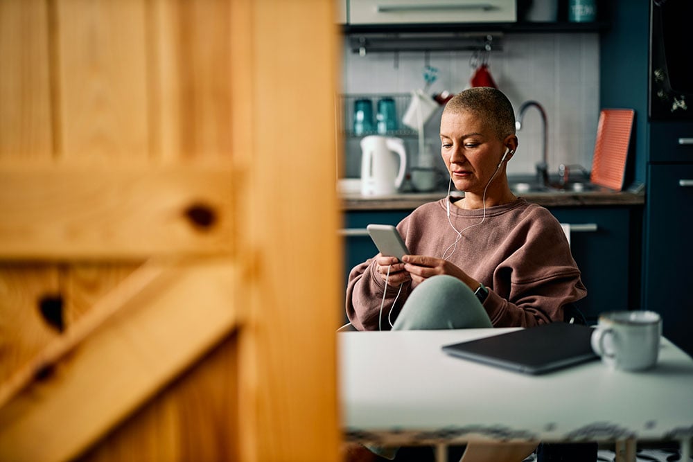 Une grand-mère (grand-parents) est assise à une table dans une cuisine moderne. Elle écoute quelque chose à travers ses écouteurs tout en regardant attentivement son smartphone. La tasse de café à côté d'elle et le carnet sur la table suggèrent une pause ou un moment de réflexion. Peut-être est-elle en train de communiquer avec ses proches ou de regarder des photos de sa famille, évoquant les défis et les émotions de la maternité à distance. Le cadre intime de la cuisine et son expression concentrée traduisent le désir de rester connectée malgré l'éloignement physique.