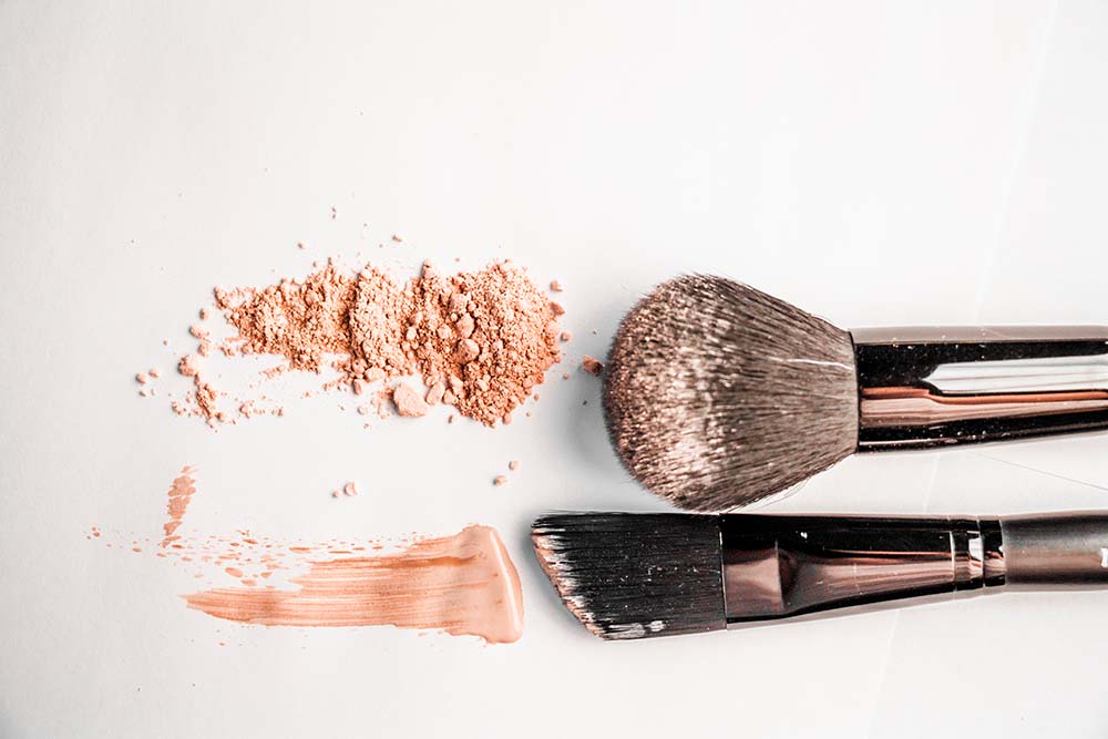 Photo mettant en scène une poudre minérale de teinte naturelle éparpillée sur une surface blanche, accompagnée de deux pinceaux de maquillage : l'un avec des poils denses pour l'application du fond de teint et l'autre plat pour les détails. Ces éléments évoquent l'utilisation de produits cosmétiques naturels et doux, spécialement conçus pour répondre aux besoins des femmes enceintes soucieuses des ingrédients qu'elles appliquent sur leur peau