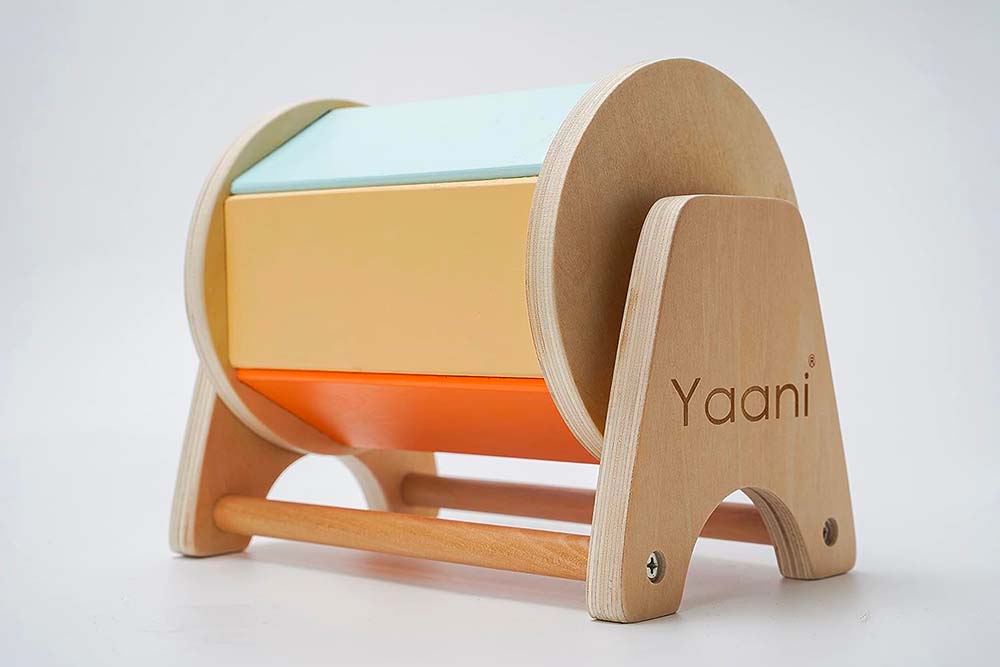 Un jouet Montessori en bois élégamment conçu pour bébé, portant la marque 'Yaani'. Le jouet présente une structure en bois naturel courbée, soutenant des blocs rectangulaires en pastel dans des teintes de turquoise, beige et orange. La conception minimaliste, avec ses lignes épurées et ses couleurs douces, est caractéristique des matériaux Montessori qui encouragent l'apprentissage autodirigé et le développement sensoriel des enfants. Le bois utilisé démontre une finition de haute qualité, avec une attention particulière aux détails, notamment les fixations discrètes et la gravure de la marque.