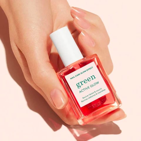 Photo d'une main délicatement posée tenant un flacon de vernis à ongles de couleur corail nommé 'green ACTIVE GLOW'. L'étiquette met en avant son effet 'NAIL CARE GLOW EFFECT' et mentionne qu'il est formulé sans toxiques. Le fond de l'image est de couleur rose pastel, évoquant un produit doux et naturel, idéal pour les femmes enceintes cherchant des cosmétiques sans danger pour le bébé et sans odeur