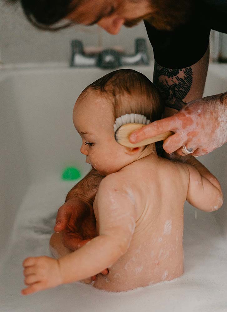 Un père, reconnaissable par ses tatouages et une barbe partielle, baigne délicatement son bébé dans la baignoire. La brosse douce qu'il utilise pour caresser la tête du nourrisson contraste avec son apparence robuste. Bien que ce soit un moment tendre, le contexte de la photo peut évoquer la lourdeur des responsabilités parentales et les défis cachés auxquels sont confrontés de nombreux pères, tels que le burn-out parental ou les sentiments associés à la dépression post-partum. La vulnérabilité de l'enfant dans cette scène amplifie l'importance du rôle du parent et les émotions potentielles qui y sont liées.