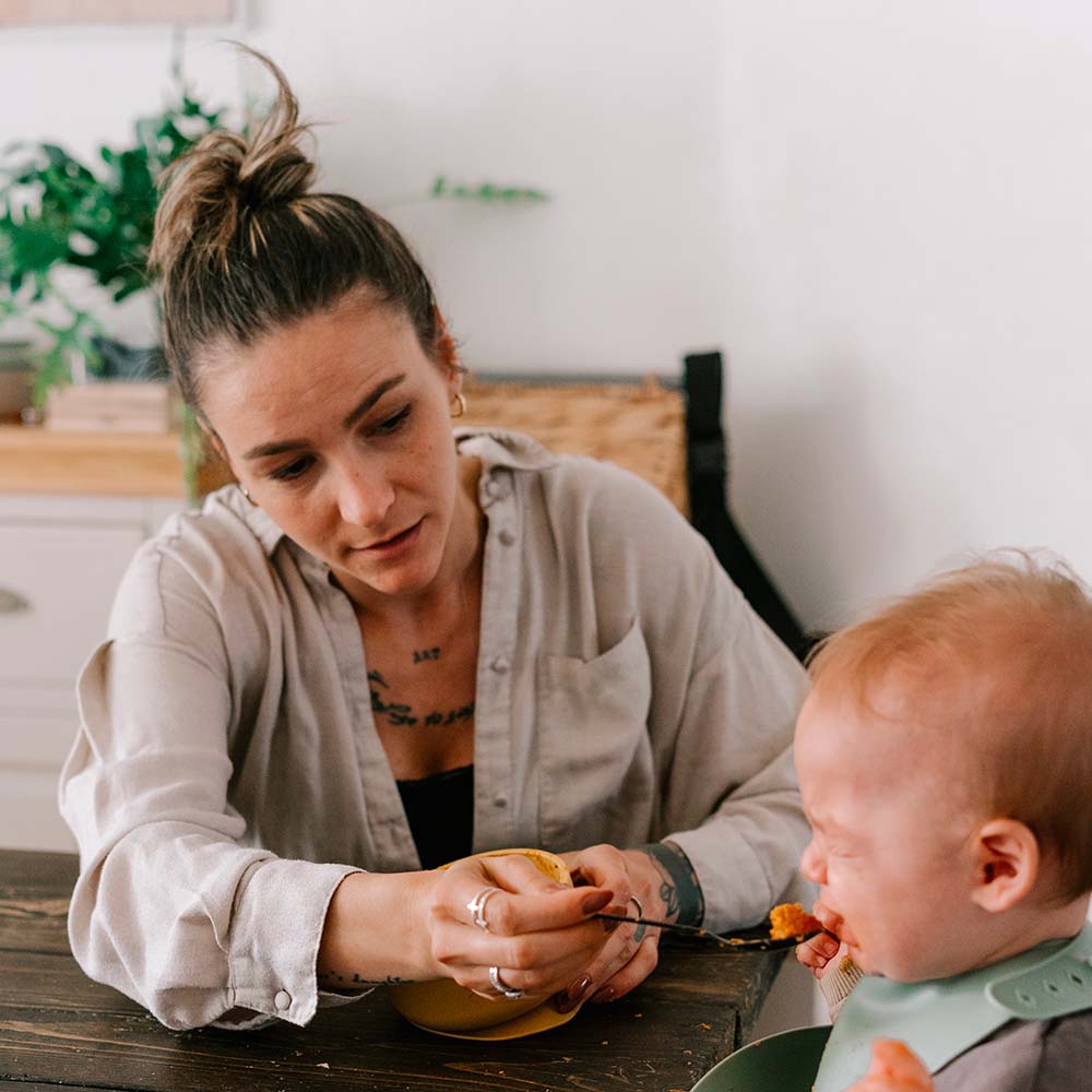 Dans un cadre domestique, une mère au regard préoccupé et aux traits fatigués donne à manger à son bébé assis à table. Alors que le bébé s'apprête à manger une cuillerée, l'expression de la mère reflète une lourdeur émotionnelle, évoquant les défis intenses de la maternité et les sentiments associés à la dépression post-partum, au baby blues et au burn-out parental. Le contraste entre la scène quotidienne et le poids des émotions maternelles est palpable.