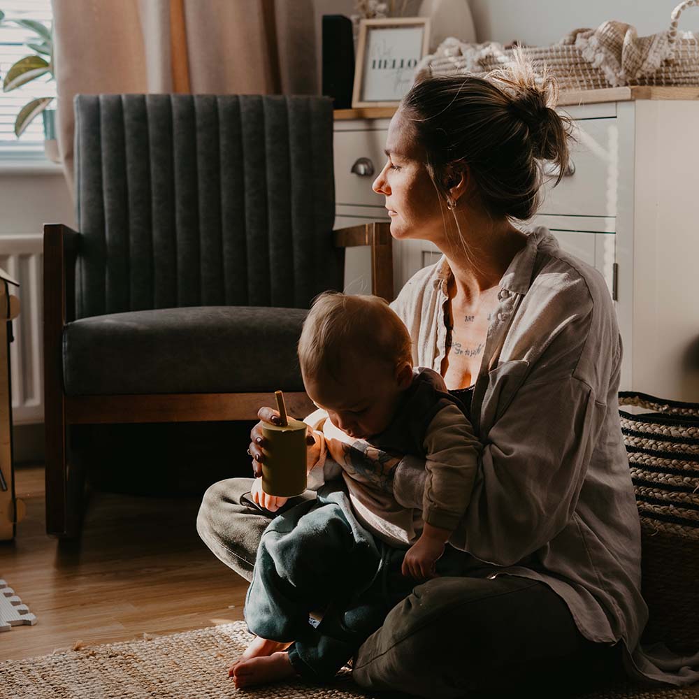 Dans une ambiance douce et éclairée par la lumière naturelle, une mère assise sur le sol étreint tendrement son bébé qui tient une tasse. Son regard lointain et sa posture introspective illustrent les défis silencieux de la maternité, évoquant les sentiments profonds liés au baby blues, à la dépression post-partum et au burn-out parental. L'environnement paisible contraste avec la lourdeur potentielle des émotions ressenties.