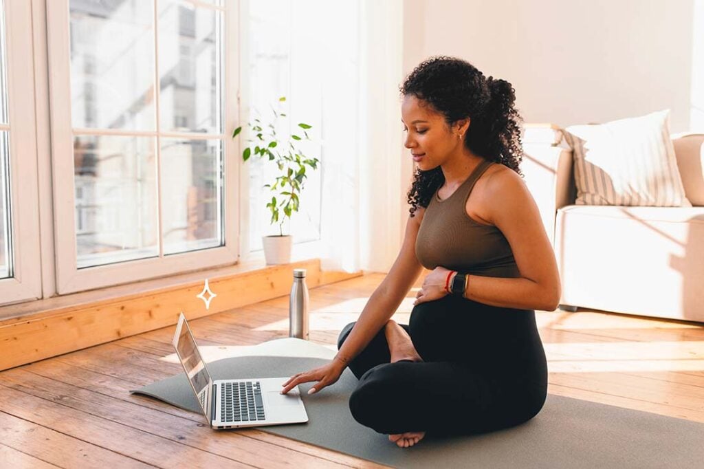 Femme enceinte confortablement assise en tailleur sur un tapis de yoga dans un salon lumineux, interagissant avec un cours de yoga prénatal en ligne sur son ordinateur portable. À ses côtés, une gourde et des écouteurs soulignent un mode de vie sain et actif. Cette scène tranquille capte l'engagement de la femme enceinte dans son bien-être et sa santé, et la technologie comme outil d'accompagnement dans sa routine d'exercice pendant la grossesse, favorisant la flexibilité et la relaxation.