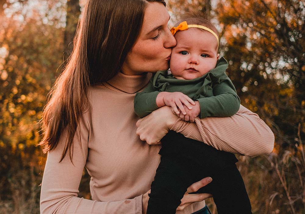 Maman embrassant affectueusement son bébé sur la joue, un exemple vivant du conseil pour créer du lien avec son bébé. La mère, en tenue décontractée, tient son enfant de manière sécurisée et aimante, tandis que le bébé, avec un bandeau jaune, regarde directement l'objectif, semblant à l'aise et en confiance dans les bras de sa mère. Atmosphère de bien-être et d'affection. Cette scène capture l'importance des gestes tendres et de l'attention portée aux jeunes enfants pour renforcer le lien d'attachement entre les parents et leur bébé.