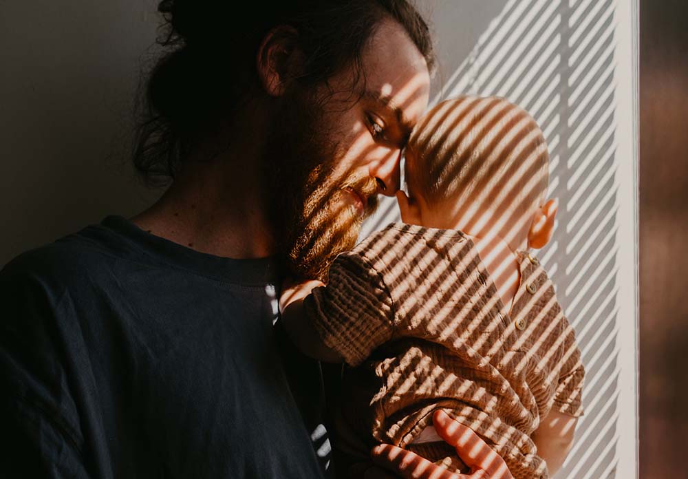 Force du "lien d'attachement bébé" entre un père et son enfant. On y voit un père barbu tenant son bébé contre lui, leurs visages touchant presque, dans un moment de complicité et d'affection. La lumière naturelle qui filtre à travers les stores crée un motif de lignes ombrées sur eux, ajoutant une dimension artistique à cette scène tendre. Ce tableau évoque l'importance du contact physique et émotionnel dans le développement d'une relation forte et sécurisante entre un parent et son bébé dès les premières étapes de la vie.