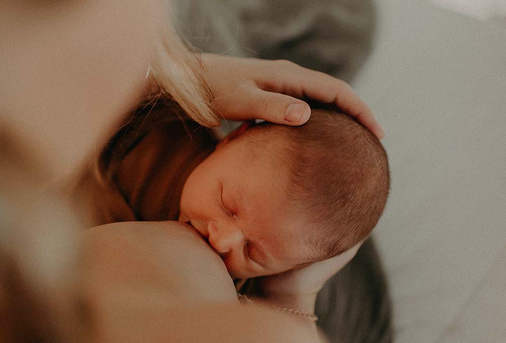 Moment intime entre une mère et son bébé, mettant en avant le concept du "lien d'attachement bébé". Le nourrisson, la tête posée confortablement contre la poitrine de sa mère, semble en pleine séance d'allaitement, un acte qui renforce le lien affectif et la sécurité émotionnelle entre l'enfant et le parent. La tendresse de la scène est soulignée par la main délicate de la mère caressant doucement la tête du bébé, illustrant le soin et l'amour inconditionnel qui caractérisent les premiers stades de l'attachement parental.