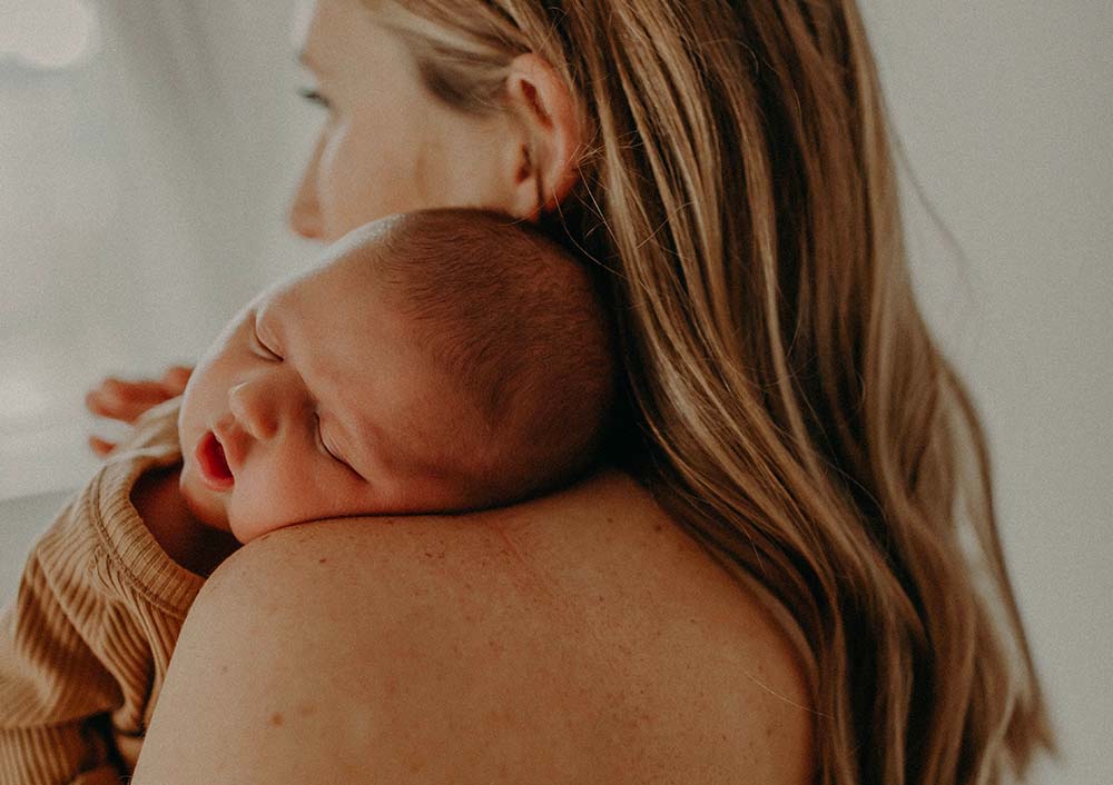 Une jeune femme tenant doucement un nouveau-né contre son épaule. Le bébé semble paisiblement endormi, blotti contre la femme dans une expression d'abandon et de confort total. Cet instant tendre illustre le thème du "lien d'attachement bébé", mettant en lumière la connexion profonde et instinctive qui se forme entre un parent et son enfant dès les premiers moments de la vie. La lumière douce et l'ambiance sereine renforcent le sentiment de sécurité et d'amour qui est essentiel au développement émotionnel d'un nourrisson.