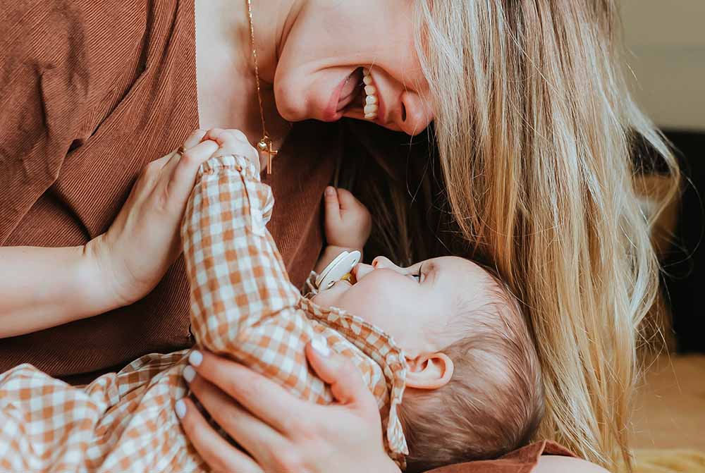 Moment joyeux et intime qui reflète le lien d'attachement affectif et psychique de bébé pendant la grossesse. Une femme souriante interagit avec un bébé allongé sur ses genoux, tenant ses petites mains. L'enfant regarde vers le haut avec confiance et intérêt, capturé dans un échange affectueux avec la mère. Leur interaction, pleine de rires et de complicité, suggère un fort sentiment de connexion et d'amour. Cette scène chaleureuse symbolise le lien émotionnel fort qui se développe entre un parent et son enfant, une base essentielle pour la confiance en soi et la sécurité émotionnelle de l'enfant dès le début de sa vie.