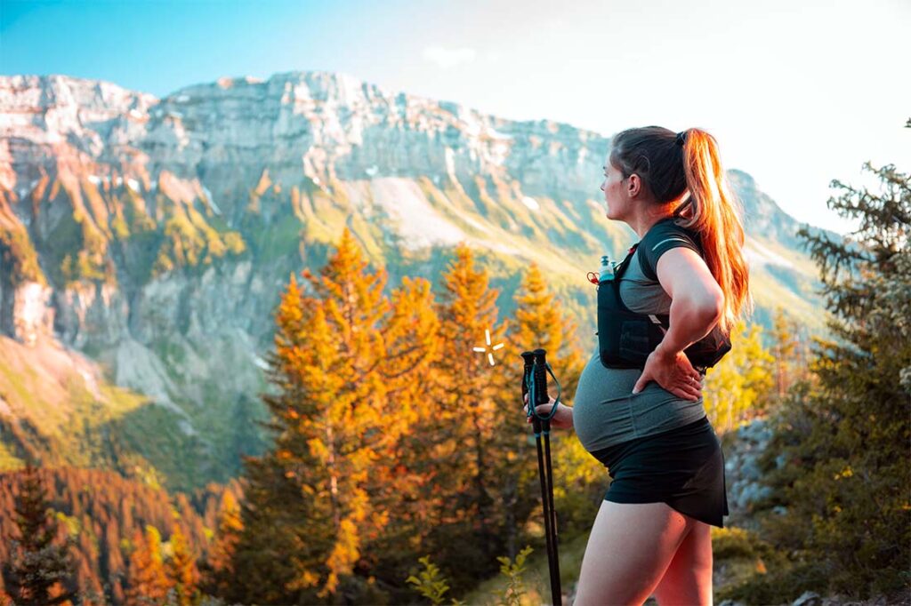 Femme enceinte effectuant une randonnée en montagne, équipée de bâtons de marche, profitant de la nature et de l'exercice physique adapté à sa grossesse. Le paysage montagneux à l'arrière-plan avec des arbres aux couleurs automnales ajoute à la beauté et au sentiment de bien-être dégagé par l'activité.