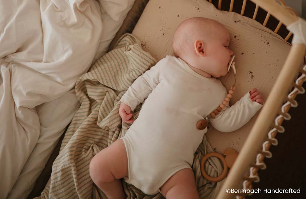 Nouveau-né dormant sereinement sur le côté dans un lit bébé, vêtu d'un body blanc à manches longues et près d'un anneau de dentition en bois avec des perles. Cet environnement apaisant est l'exemple d'une routine de sommeil réglée par la méthode 'chrono-dodo', conçue pour aider les bébés à s'endormir de manière régulière et paisible.