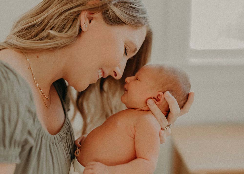 Lien d'attachement bébé avec sa mère et son enfant en interaction face à face. La mère, souriante et visiblement émue, se penche vers son bébé qui la regarde attentivement. Le contact visuel direct et l'expression faciale du bébé suggèrent une reconnaissance et un confort émotionnel, des composantes clés du développement d'un lien d'attachement sécurisé. La chaleur et l'affection palpable entre la mère et son enfant sont capturées dans cette image douce et lumineuse, reflétant l'importance des premières interactions pour établir un lien parental fort.