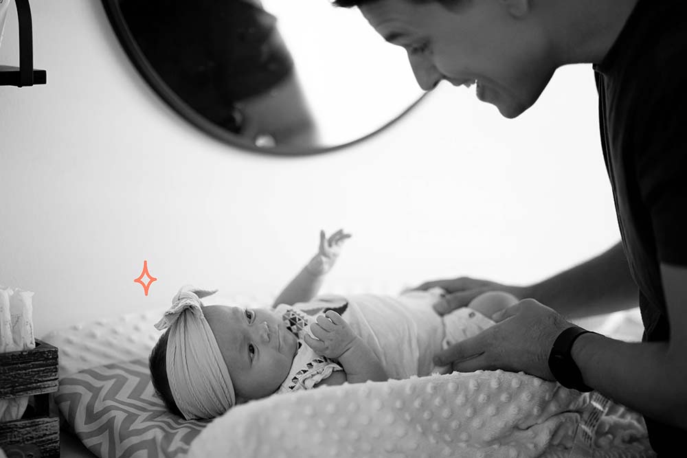 Un père jouant et interagissant avec son bébé allongé sur un tapis à motifs, avec son reflet dans le miroir, illustrant l'importance de l'engagement paternel et de la connexion émotionnelle pendant la période d'allaitement.