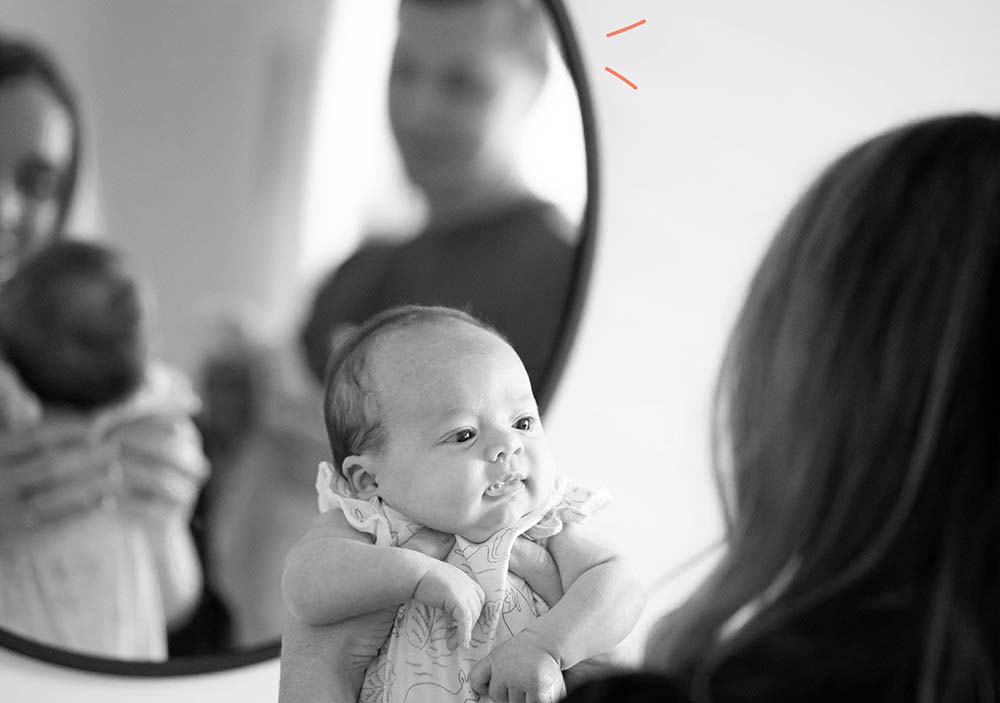 Un père souriant reflété dans un miroir tenant son bébé, tandis que la mère allaite en arrière-plan, symbolisant la participation active du père dans les soins du nourrisson et son rôle de soutien durant l'allaitement.