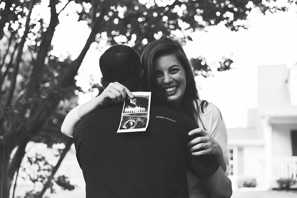 Un couple heureux en extérieur, l'homme de dos avec une échographie du premier trimestre après le 1er rendez-vous prénatal, représentant la joie et le partage des premiers moments de la grossesse.