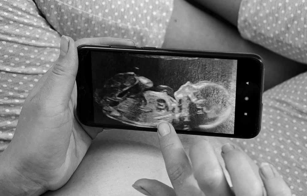 Une future mère regarde une échographie 3D de son bébé sur un smartphone, mettant en avant l'importance des rendez-vous médicaux du troisième trimestre de grossesse jusqu'aux deux ans de l'enfant.