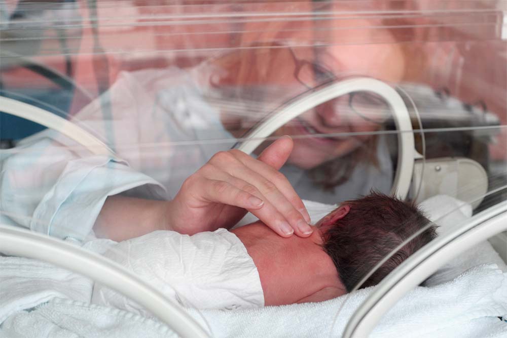 Une mère touche doucement son enfant prématuré à travers l'ouverture d'une couveuse, une image qui évoque les soins maternels intensifs et l'amour inconditionnel dans un environnement de soins néonatals pour les prématurés.