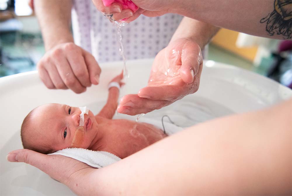Un bébé prématuré reçoit son premier bain avec précaution et amour par des mains attentives, une étape clé dans les soins post-nataux apportés aux prématurés pour assurer leur bien-être et hygiène dans une maternité.