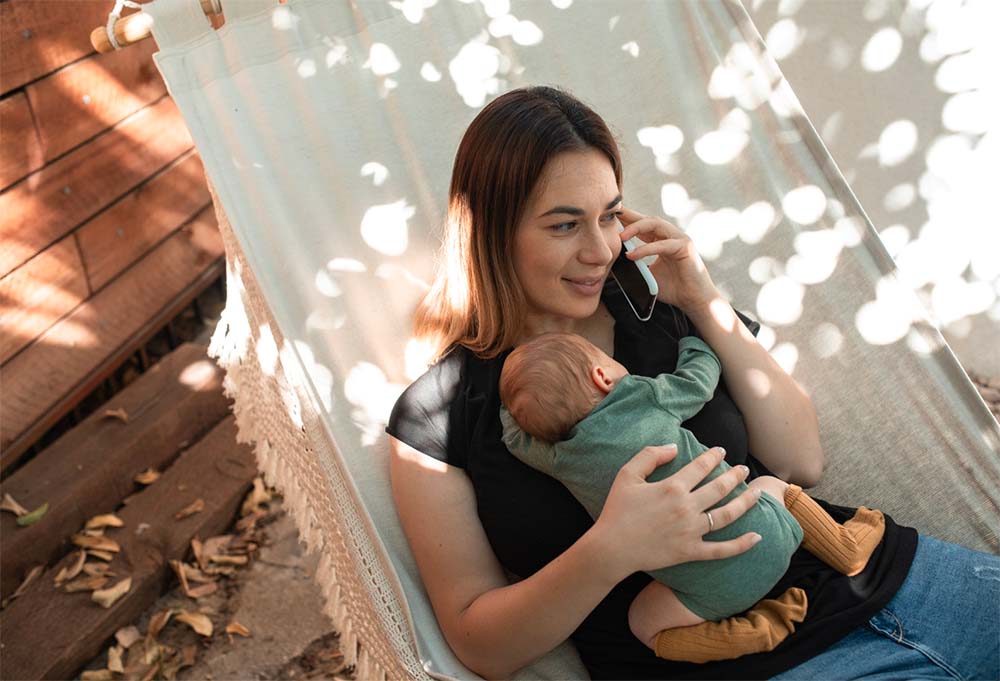 Mère multitâche tenant son bébé tout en parlant au téléphone, démarche qui reflète les étapes pour déclarer une assistante maternelle. Suivez notre tutoriel pour une embauche sereine et conforme.