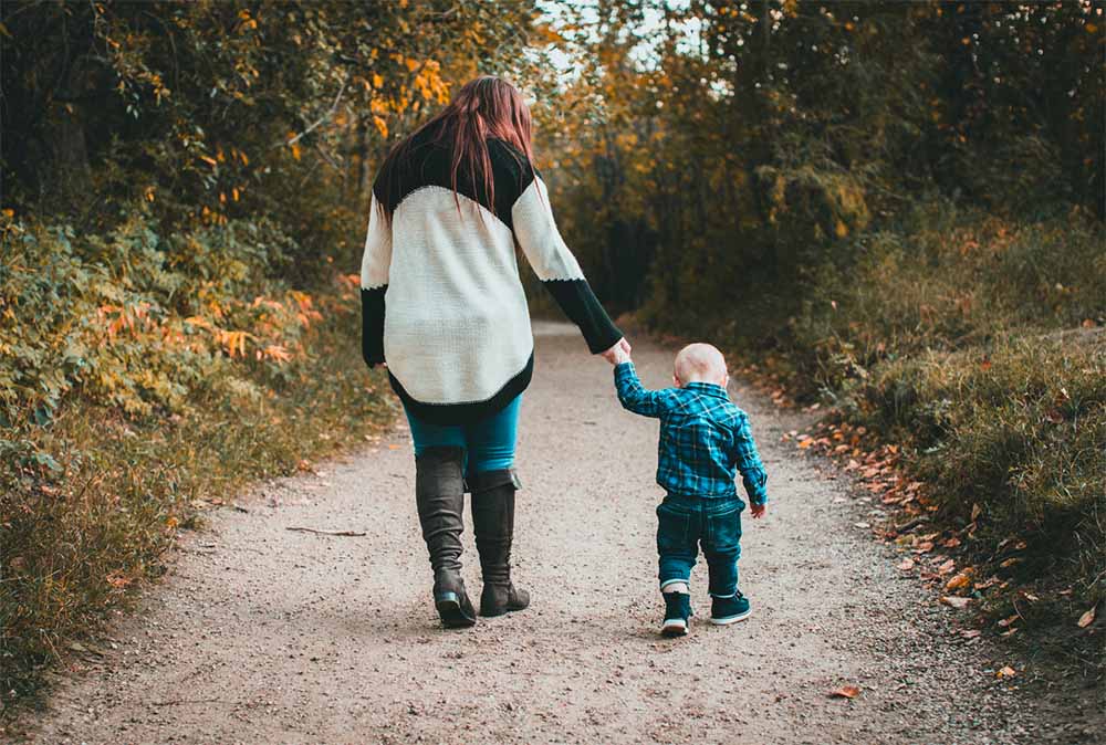 Jeune enfant marchant main dans la main avec un adulte dans un chemin boisé, évoquant la guidance et la sécurité apportées par les professionnels de la petite enfance. Guide pas à pas pour embaucher et déclarer une assistante maternelle.