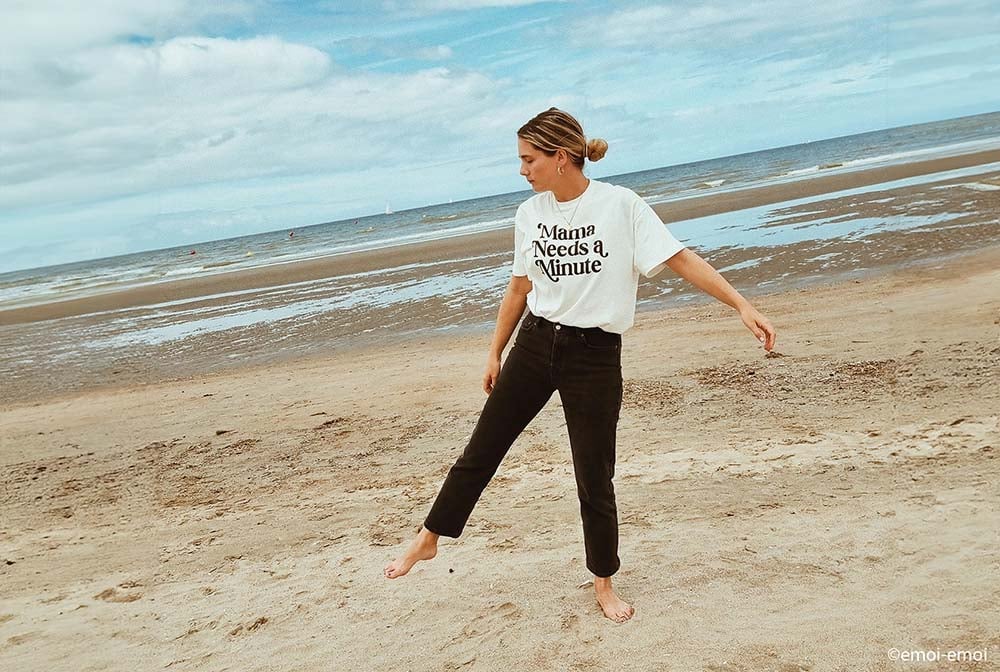 Femme enceinte décontractée sur la plage, arborant un t-shirt humoristique 'Mama Needs A Minute', assorti à un pantalon noir, illustrant un style maternité à la fois tendance et confortable pour les futures mamans.