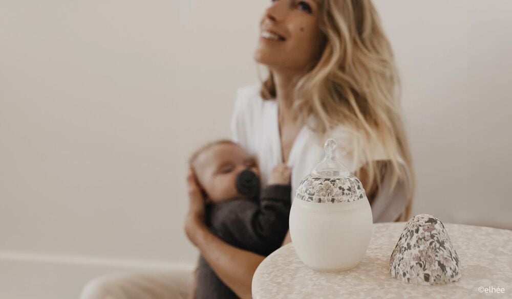 Biberon de lait maternel au design floral posé sur une table, avec une mère souriante tenant son bébé en arrière-plan