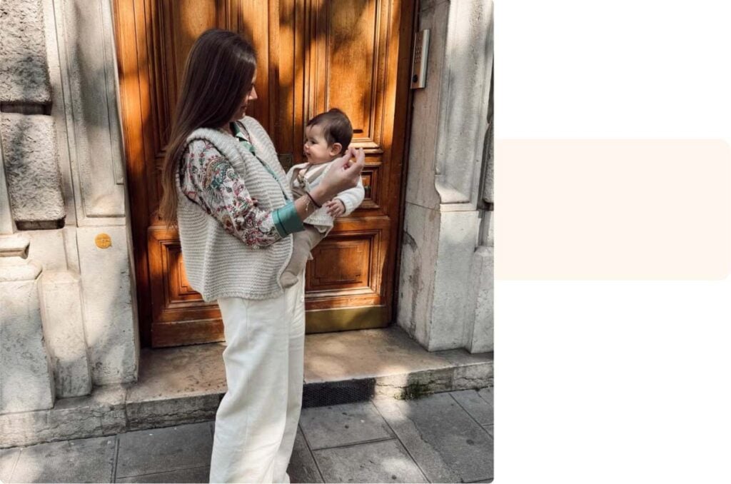 Maman souriante tenant son bébé dans les bras devant une porte ancienne, témoignage maman plein de complicité et de tendresse
