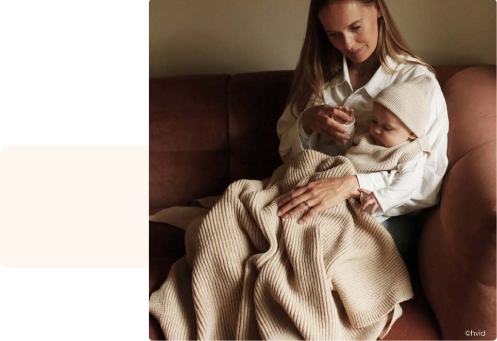 Jeune femme assise tenant son bébé dans une couverture en laine chaude, moment de tendresse pour protéger bébé du froid à la maison