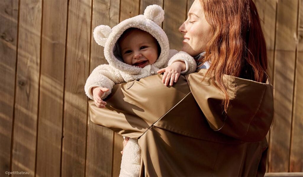 Mère souriante portant son bébé vêtu d’une combinaison à oreilles d’ourson, bien couvert pour protéger bébé du froid