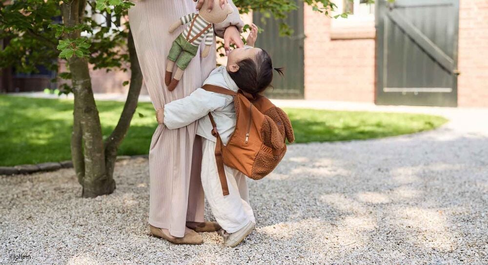 Période d’adaptation crèche bébé, enfant accroché à sa mère avec un sac à dos en peluche avant la séparation du matin.