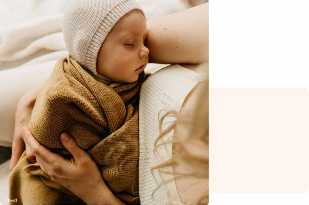 Bébé endormi dans les bras de sa mère, emmitouflé dans une couverture moutarde pour protéger bébé du froid
