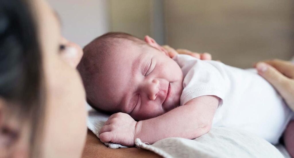 Nouveau-né souriant dans les bras de sa mère, symbole du lien renforcé grâce au soutien d’une sage-femme post-partum