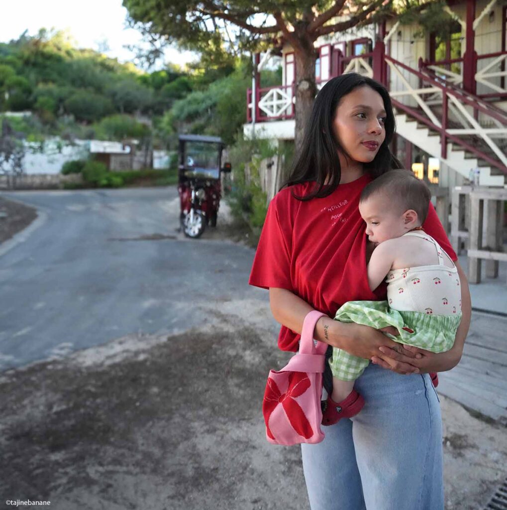 Mère en t-shirt rouge portant son bébé dans les bras devant une maison coloniale, moment d'attachement et d'allaitement maternel