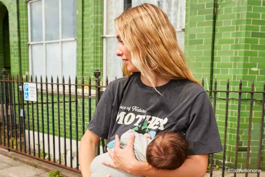 Une femme aux longs cheveux blonds porte son nouveau-né près de son cœur, dans un environnement urbain. Son regard est dirigé vers le lointain, peut-être en pensant à sa famille éloignée. Sa chemise noire, imprimée du mot "MOTHER" (mère), symbolise son rôle récent et sa force. Bien qu'elle soit physiquement éloignée de sa famille d'origine, le lien intime et protecteur qu'elle entretient avec son bébé est évident. L'arrière-plan urbain et les briques vertes mettent en avant le contraste entre la douceur du moment maternel et le tumulte de la ville. Avoir un bébé loin de sa famille peut être un défi, mais cette image montre la beauté et la résilience de la maternité malgré la distance.