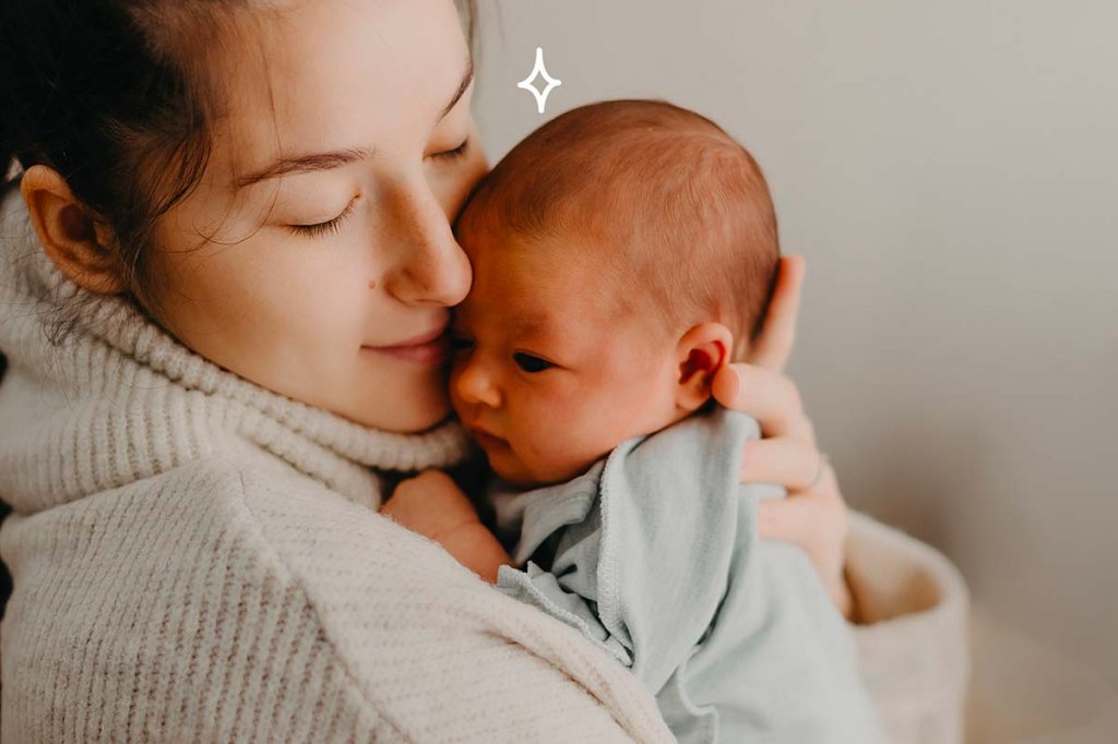 Jeune mère tenant son bébé contre elle dans un tendre câlin, incarnant le "lien d'attachement bébé". La mère, les yeux fermés et un sourire serein sur le visage, semble savourer le moment avec son enfant. Le bébé, regardant attentivement sa mère, est enveloppé dans une couverture douce, ce qui suggère chaleur et sécurité. Cette scène capture la connexion émotionnelle profonde et l'importance des premiers contacts physiques qui sont essentiels au développement d'un lien sûr et aimant entre un parent et son enfant.