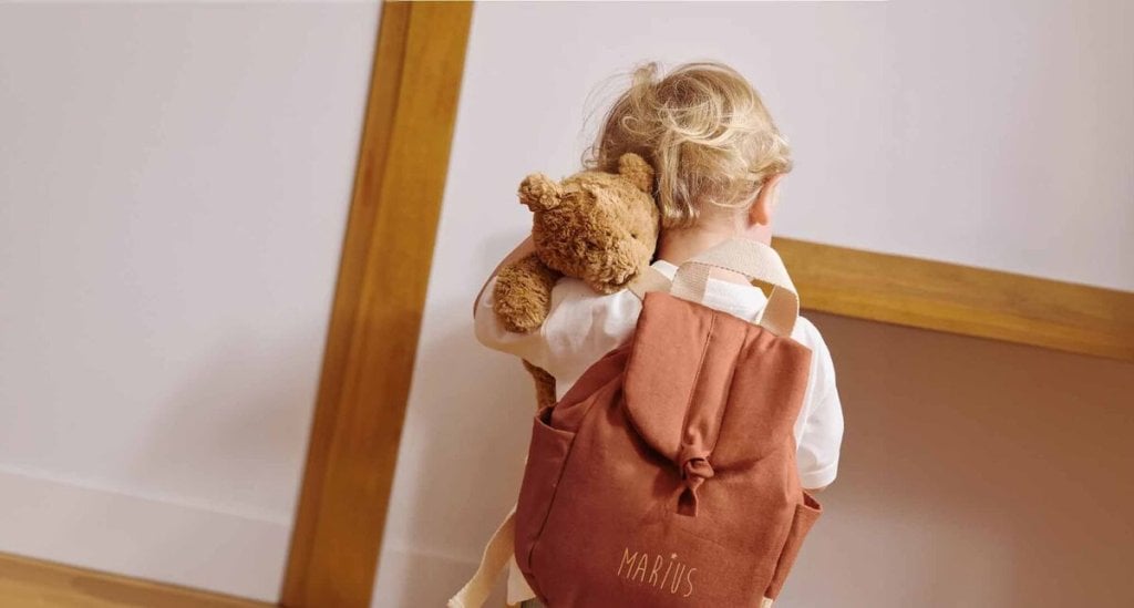 Bébé de dos portant un sac à dos et tenant un ours en peluche, illustrant la période d’adaptation à la crèche pour bébé