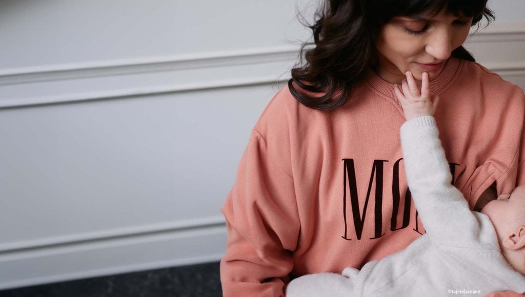 Mère portant un sweat rose avec l'inscription "MOM" allaite son bébé en extérieur
