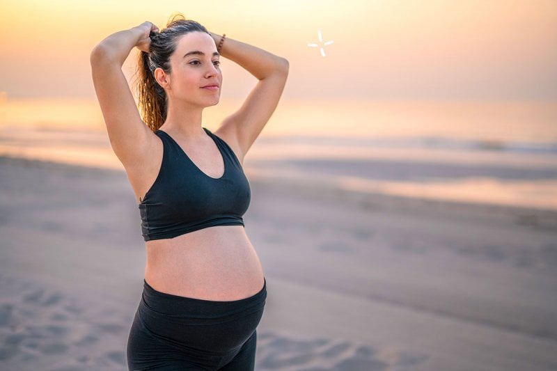 Femme enceinte souriante faisant des exercices de fitness sur une plage au coucher du soleil, démontrant l'importance du sport et de l'activité physique pendant la grossesse pour le bien-être et la santé.