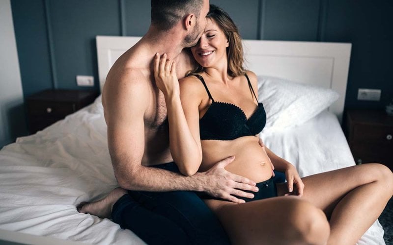 Couple enceinte partageant un moment intime et affectueux sur un lit, symbolisant la connexion et le sexe pendant la grossesse dans une chambre paisible. La femme enceinte, souriante et confortablement installée, reçoit des caresses de son partenaire, ce qui souligne l'importance de l'intimité et de l'adaptation sexuelle pendant la grossesse.