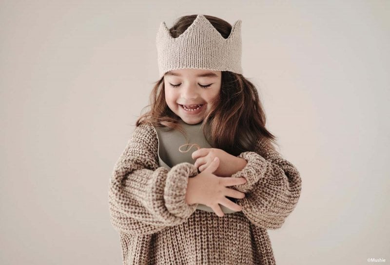 Une petite fille souriante avec une couronne tricotée, évoquant la douceur et l'originalité pour inspirer le choix d'un prénom rare et cool pour une fille. Liste de prénoms.